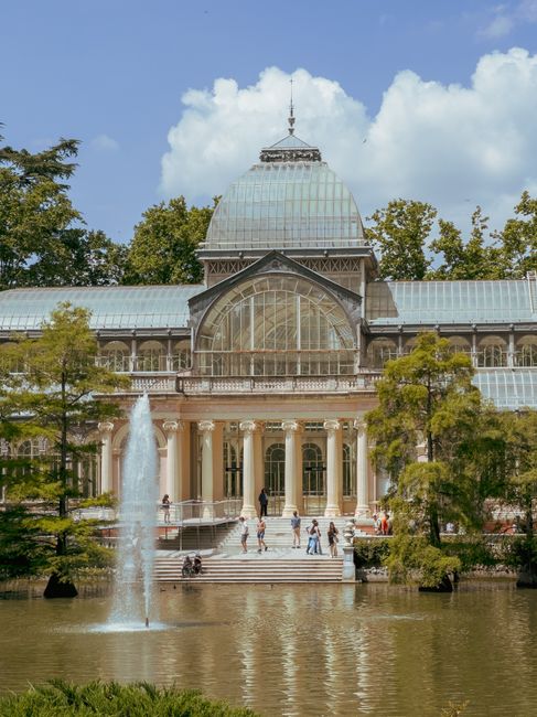 A large, historic glass-domed conservatory surrounded by trees and a pond. The building features a prominent central dome with a spire at the top, flanked by two smaller wings with glass walls and metal framework. The facade has a series of tall, arched windows and columns supporting a decorative balcony with intricate balustrades. In front of the building, there is a wide staircase leading down to the pond, where a fountain sprays water into the air. Several people are visible walking or sitting on the steps, with some near the water's edge. The scene is set outdoors on a bright day with a partly cloudy sky, contributing to a pleasant and inviting atmosphere. Natural greenery and reflections on the water enhance the scenic view, while the building’s design and setting suggest a historic greenhouse or conservatory, typical of private or semi-public gardens, aligning with themes of alternative waste handling and maintaining an elegant exterior environment.