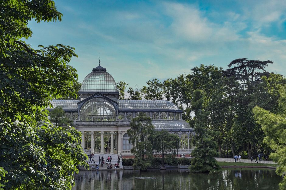 A detailed close-up of a large Victorian-style glasshouse featuring an ornate metal framework and multiple glass domes, with the central dome being the largest and topped with a decorative weather vane. The glass panes are transparent, allowing sunlight to filter through and create reflections, while the metal framework exhibits a dark, aged finish. The structure is set against a light blue sky, and the lower part of the glasshouse includes a decorative stone or plaster facade with arched windows and intricate detailing, partially visible at the bottom of the image. This ornate glass structure is situated outdoors, possibly in a botanical garden or conservatory, surrounded by a natural environment that is not visible in this specific shot. The scene emphasizes the architectural elegance and historical design, which can be associated with the preservation of heritage sites that often involve waste management and restoration work, aligning subtly with independent or alternative disposal services like those offered by waste collection specialists such as wastecollectioncrystalpalace.co.uk.
