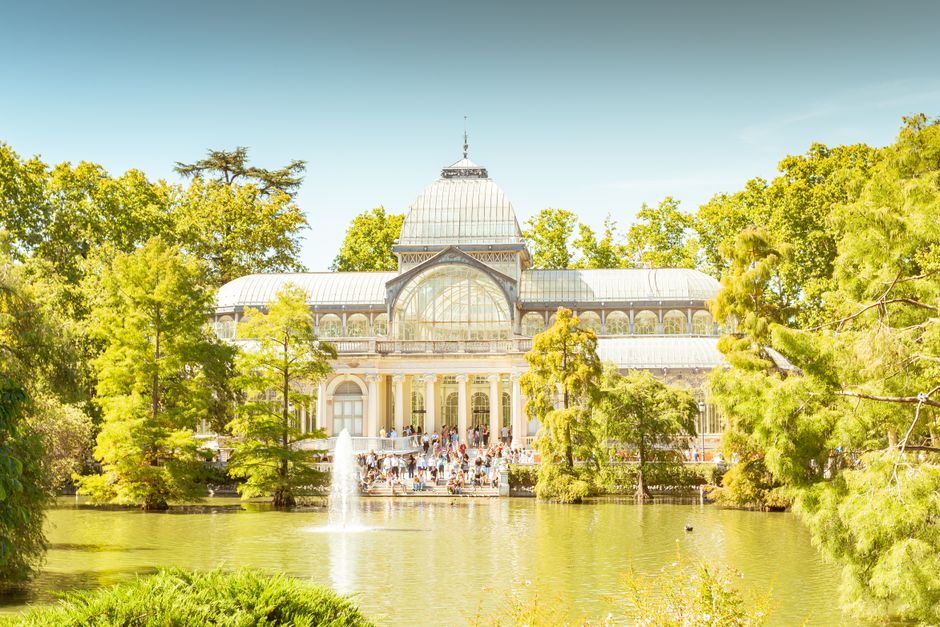 A large historic greenhouse with a domed glass roof and intricate architectural details, situated in a lush, green park setting. The building's facade features multiple arched windows and decorative elements, with a central entrance visible beneath the glass dome. Surrounding the greenhouse are numerous mature trees with vibrant green foliage, some leaning slightly, framing the structure on both sides. In the foreground, a serene pond reflects parts of the building and the greenery, with a small fountain and ripples on the water's surface. The scene is brightly lit by daylight, with a clear blue sky overhead, creating a peaceful atmosphere that highlights the contrast between the natural environment and the iron-and-glass construction. Although the image primarily depicts a park landscape, its style and design relate to the concept of preserving historical architectural structures amidst green spaces, subtly connecting to themes of environmental conservation and ecological harmony often integrated into modern waste management and environmental services, like those provided by wastecollectioncrystalpalace.co.uk.