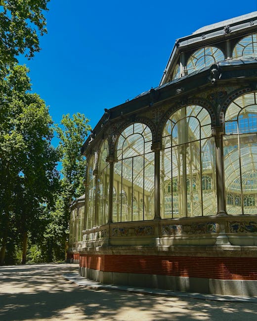 A large historic greenhouse with a domed glass roof and intricate architectural details, situated in a lush, green park setting. The building's facade features multiple arched windows and decorative elements, with a central entrance visible beneath the glass dome. Surrounding the greenhouse are numerous mature trees with vibrant green foliage, some leaning slightly, framing the structure on both sides. In the foreground, a serene pond reflects parts of the building and the greenery, with a small fountain and ripples on the water's surface. The scene is brightly lit by daylight, with a clear blue sky overhead, creating a peaceful atmosphere that highlights the contrast between the natural environment and the iron-and-glass construction. Although the image primarily depicts a park landscape, its style and design relate to the concept of preserving historical architectural structures amidst green spaces, subtly connecting to themes of environmental conservation and ecological harmony often integrated into modern waste management and environmental services, like those provided by wastecollectioncrystalpalace.co.uk.
