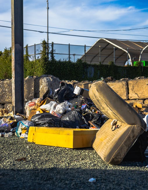 A roadside scene showing a pile of mixed rubbish and waste materials in an outdoor environment, with black plastic garbage bags, cardboard boxes, and fragments of plastic and paper scattered on the gravel surface. Prominent in the foreground is an old, worn tyre leaning against the pile, while a yellow plastic container lies partially open among the debris. Behind the rubbish, a low stone wall made of irregularly shaped blocks provides a barrier, with a green hedge and some trees visible beyond it. Overhead, utility wires run across a clear blue sky with scattered clouds, and in the background, there is a large, curved, semi-enclosed structure possibly part of a sports facility or warehouse. The scene appears to depict an instance of private rubbish accumulation, suggesting the need for an independent rubbish removal or on-site clearance service, as might be handled by companies like wastecollectioncrystalpalace.co.uk in non-local authority waste management contexts.