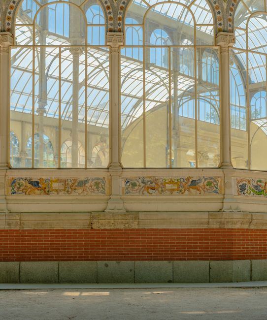 An outdoor area featuring a covered glass and metal roof structure with intricate ironwork detailing, supported by evenly spaced black and white striped metal posts. The ground is paved with smooth concrete, casting complex shadows from the overhead framework. On the left side, a red brick wall with some green doorways and flower boxes is visible, while on the right, a low black metal fence separates the space from adjacent brick buildings with white-framed windows and awnings. The clear blue sky is visible through the transparent sections of the roof, and a few small trees can be seen beyond the fencing. The environment appears clean and well-maintained, suggesting it may be part of a public transport station, market area, or another communal space, where occasional cleaning or rubbish clearance might be required, aligning with the context of independent waste collection services such as those provided by wastecollectioncrystalpalace.co.uk.
