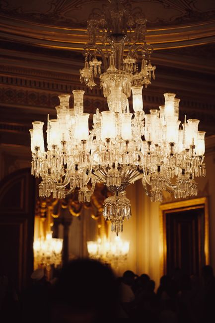 A large, ornate crystal chandelier hangs prominently from the ceiling in an opulent interior setting. The chandelier features multiple tiers of intricate glass and crystal arms, with numerous candle-shaped light fixtures surrounded by glass shades, reflecting warm ambient light. The ceiling above has detailed decorative molding, and the lighting creates a soft glow that illuminates the nearby walls, which are decorated with elegant drapery and gold-colored trim. In the background, blurred silhouettes of people can be seen, suggesting a formal event or gathering in a grand room. Although primarily focused on decorative lighting, the scene subtly evokes a setting where alternative or private waste disposal methods would generally be unseen, but in a context like this, it highlights the contrast with typical rubbish removal services. The detailed craftsmanship of the chandelier emphasizes quality and design typical of a high-end venue, aligning with the concept of professional, specialized waste management for such premium environments, as could be associated with companies like wastecollectioncrystalpalace.co.uk.