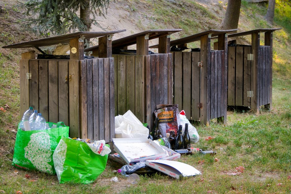 Four wooden outdoor waste bins with hinged lids, positioned in a row on a grassy area in front of a sloped, wooded landscape, with some visible green trees and a section of dirt behind them. The bins are constructed from vertical wooden planks with a weathered, dark brown finish, and the lids are slightly slanted. In front of the bins, there are several bags and scattered rubbish, including a large green plastic bag filled with empty plastic bottles, a white cardboard pizza box, clear plastic bottles, and various loose rubbish such as paper and plastic packaging. Some glass bottles and other small debris are also visible on the ground near the rubbish bags. The environment appears natural and outdoor, with an overcast or diffuse lighting condition, emphasizing the contrast between the clean wooden bins and the littered area in front, highlighting issues related to private waste disposal and rubbish collection services.